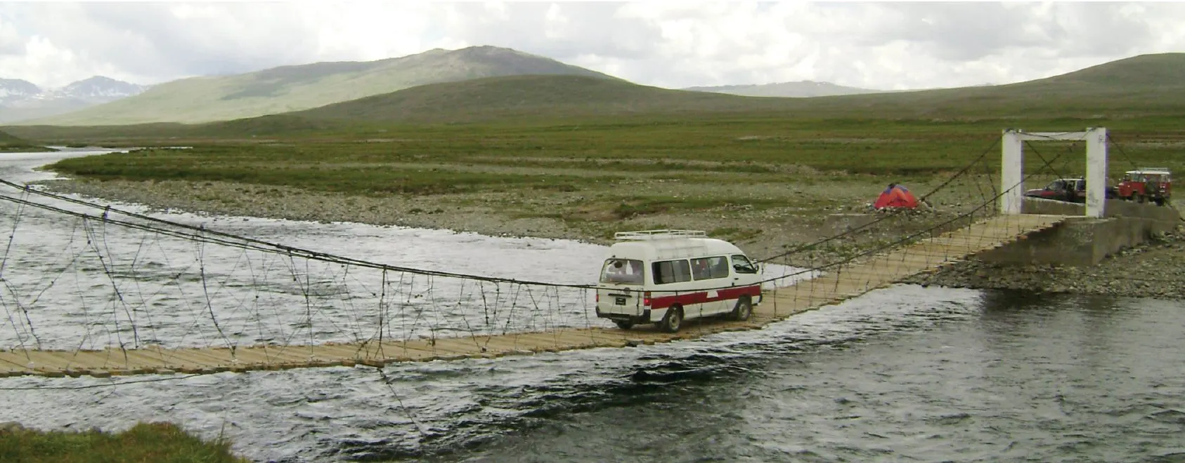 An Amber Tours van crossing a rope suspension bridge over a river on the Deosai plains