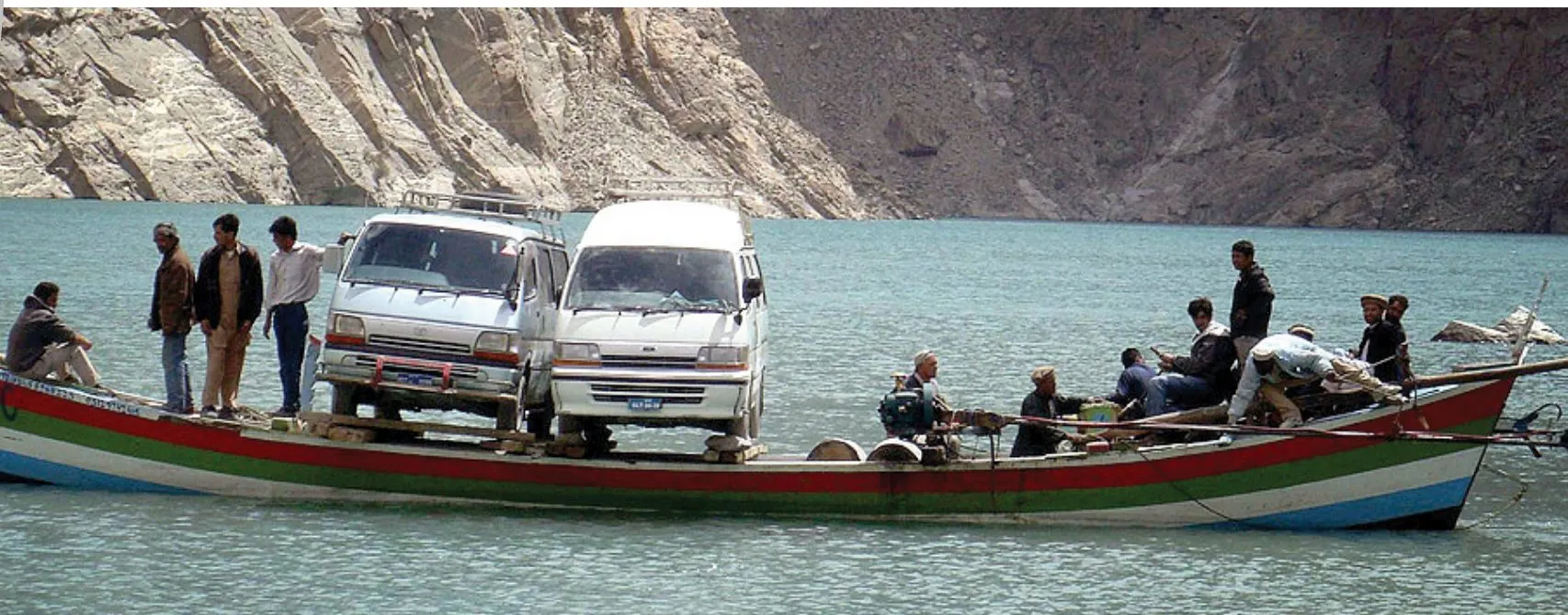 Two Hiace vans being carried across a mountain lake on a wooden boat, with passengers standing beside them