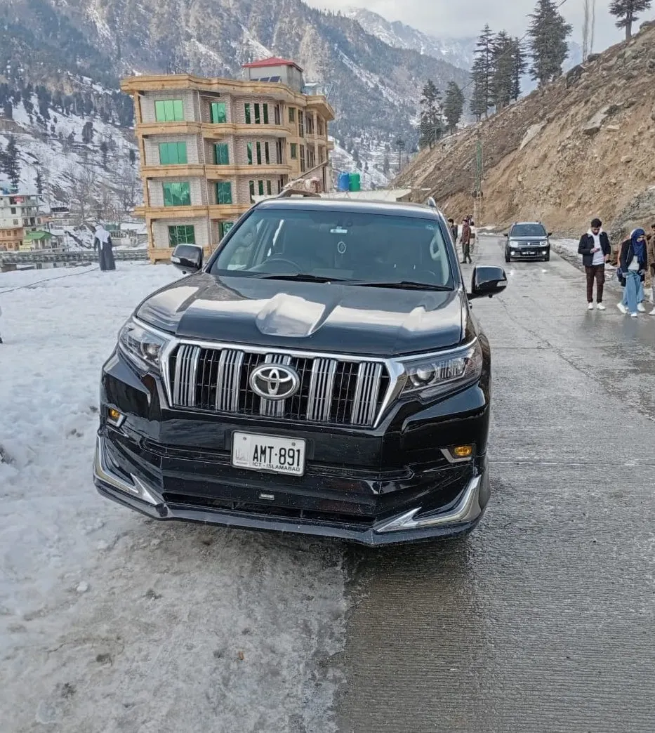 A black Toyota Prado on a snowy mountain road with a guesthouse and peaks behind